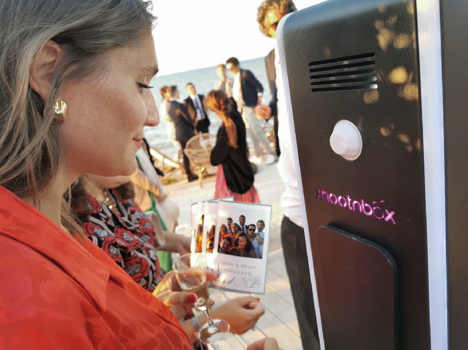 femme blonde habillé en rouge tient un verre de champagne et observe borne photo booth shootnbox logo photo imprimée