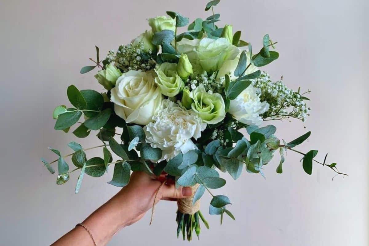 main de femme tenant un Bouquet de mariée champêtre rond en tiges naturelles Fleurs : Lisianthus vert, Oeillets blanc, Roses blanches « White naomi », Gypsophile Feuillage : Eucalyptus « Cinerea »