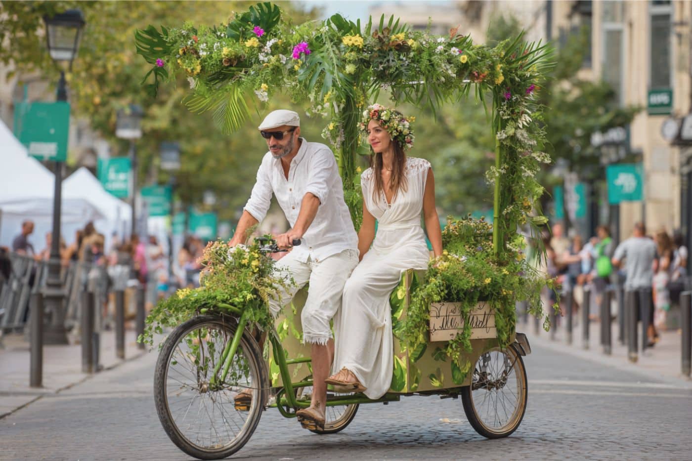 Jeune couple de mariés partant à vélo cargo décoré de fleurs locales, symbole de mobilité douce et d’un mariage éco-responsable et joyeux arrivée mariés à la mairie