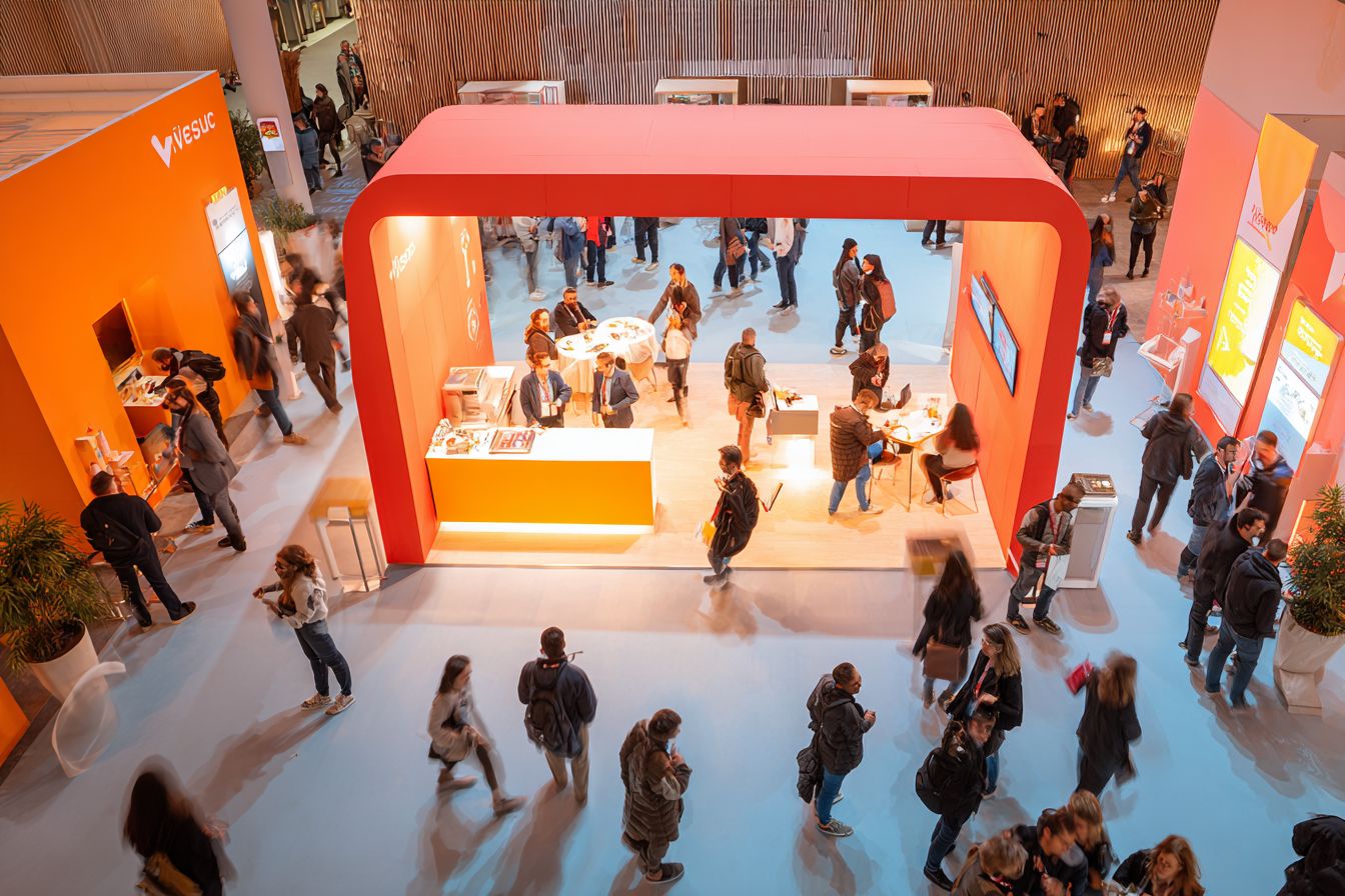 Vue du dessus d’un stand sur un salon professionnel avec photobooth lumineux, visiteurs en interaction et ambiance dynamique
